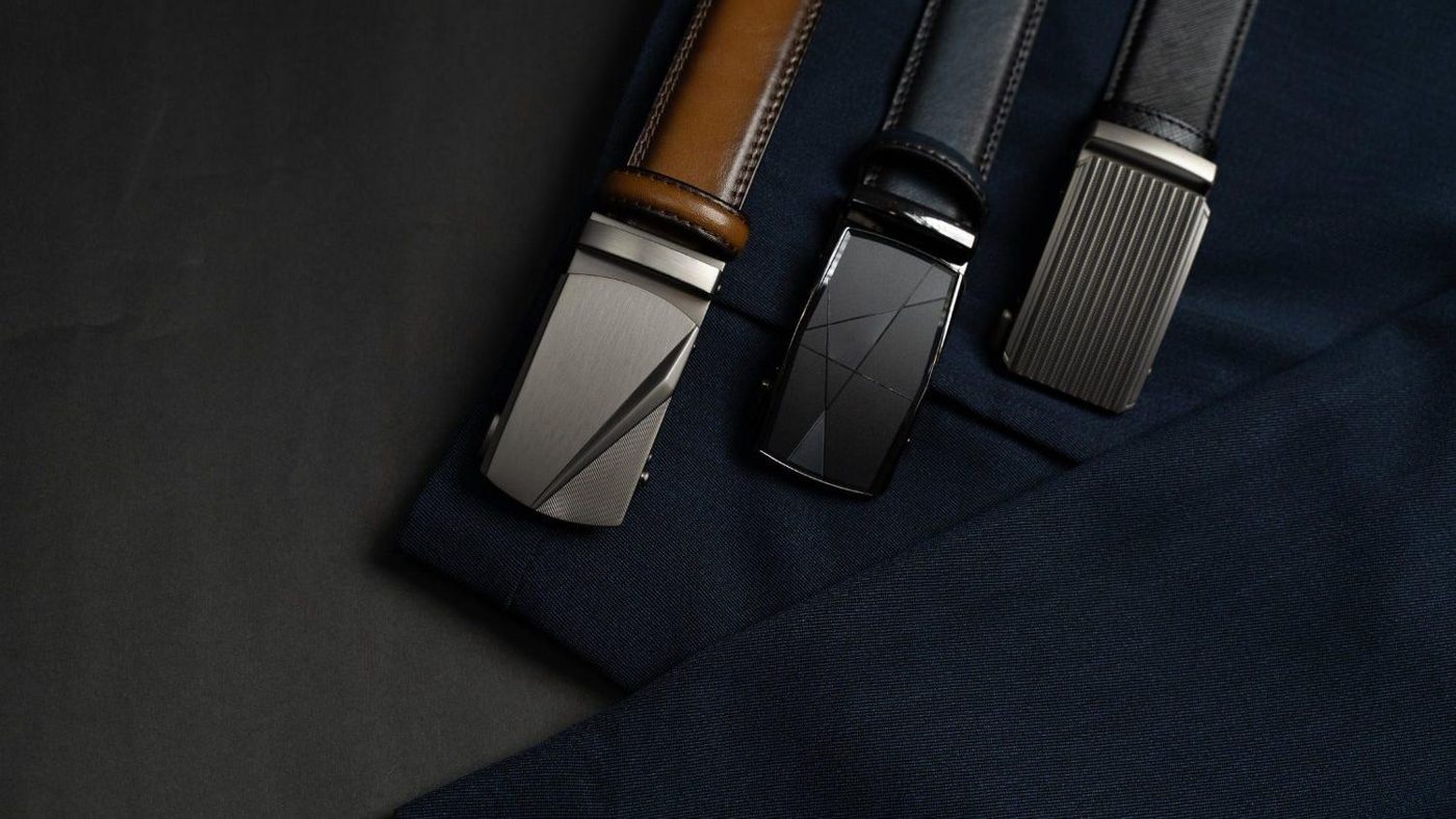 Black leather ratchet belts with engraved buckles, laid on navy fabric